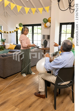Smiling woman holding gift while man with balloons points in living room 135676238