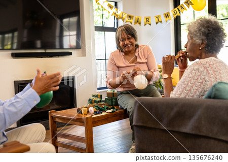 Smiling senior women celebrating birthday at home, inflating balloons with friends Smiling senior women celebrating birthday at home, inflating balloons with friends 135676240