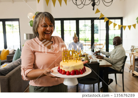 Woman carrying birthday cake with candles, smiling during celebration at home 135676289