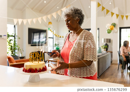 Elderly woman lighting candles on 60th birthday cake in living room celebration Elderly woman lighting candles on 60th birthday cake in living room celebration 135676299