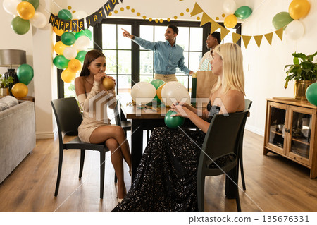 Friends celebrating birthday at home, decorating with balloons and laughing together Friends celebrating birthday at home, decorating with balloons and laughing together 135676331