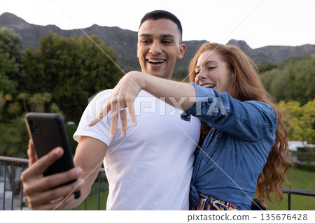 Young couple taking selfie outdoors, showing engagement ring with joyful expressions 135676428