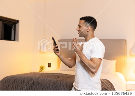Man in bedroom video chatting on smartphone, smiling and gesturing happily Man in bedroom video chatting on smartphone, smiling and gesturing happily 135676429