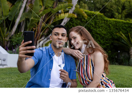 Taking selfie with champagne glasses, couple celebrating outdoors in garden 135676434