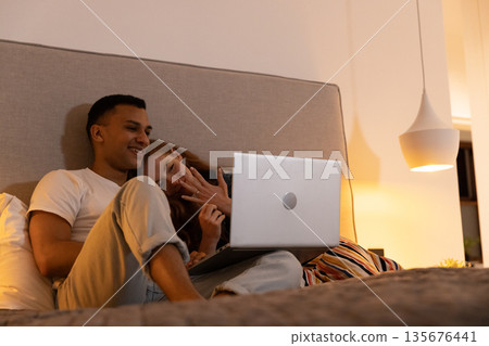Young couple relaxing in bed, watching laptop together, enjoying cozy evening Young couple relaxing in bed, watching laptop together, enjoying cozy evening 135676441