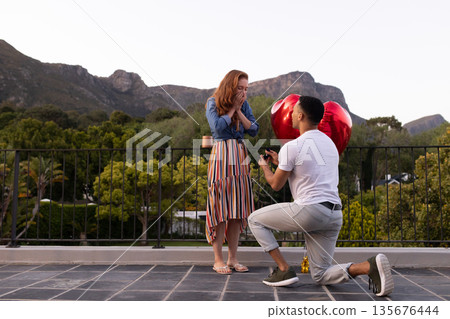 Man proposing to surprised woman with heart-shaped balloons on scenic rooftop 135676444