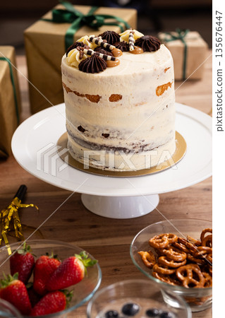 Tall cake with chocolate decorations on table surrounded by gifts and snacks 135676447