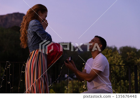 Young man proposing with ring to surprised woman on romantic evening balcony Young man proposing with ring to surprised woman on romantic evening balcony 135676448