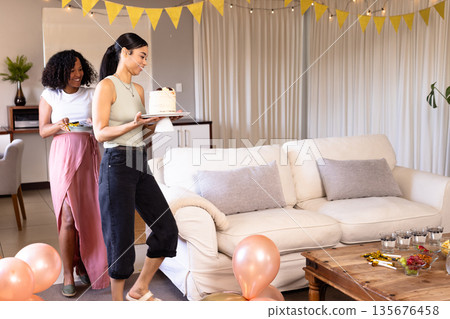 Women carrying cake and plates, preparing for celebration in cozy living room Women carrying cake and plates, preparing for celebration in cozy living room 135676458