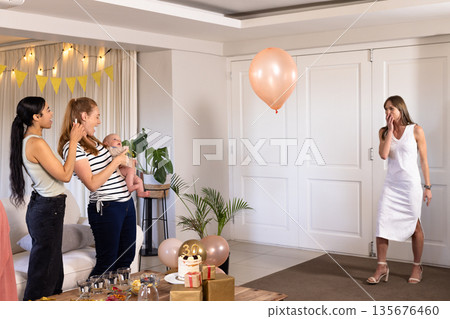 Women celebrating at home with balloons and cake, enjoying joyful moments together 135676460
