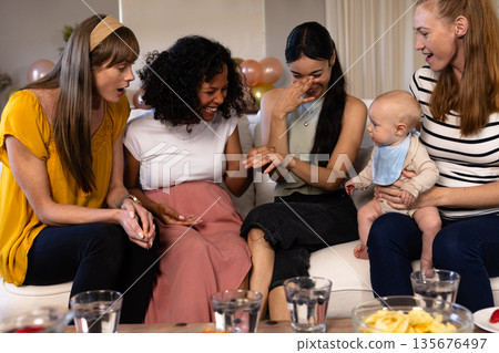 Women celebrating engagement, admiring ring, sitting on couch with baby nearby 135676497