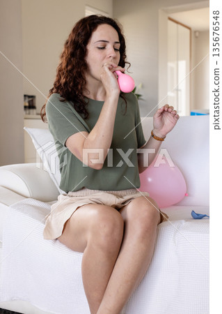 Inflating pink balloon, woman sitting on couch at home, preparing for celebration 135676548