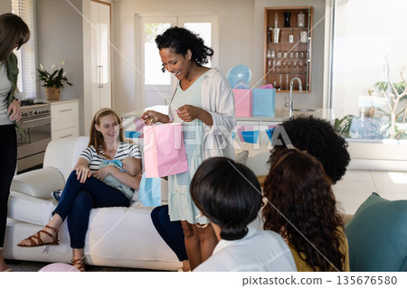 Woman opening gift bag at home, surrounded by friends during joyful celebration Woman opening gift bag at home, surrounded by friends during joyful celebration 135676580