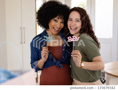 Smiling women celebrating baby shower at home, holding decorative signs Smiling women celebrating baby shower at home, holding decorative signs 135676582