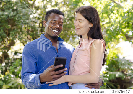 Couple enjoying outdoor moment, smiling while looking at smartphone in garden Couple enjoying outdoor moment, smiling while looking at smartphone in garden 135676647