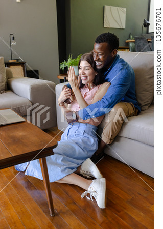 Couple sitting on floor, smiling and waving during video call at home 135676701