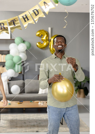 Smiling man holding gold balloon celebrating birthday at home with decorations 135676702