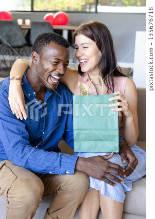 Couple celebrating with gift bag, laughing together on cozy living room sofa 135676718