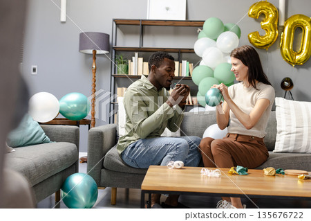 Celebrating birthday, diverse couple inflating balloons in cozy living room setting Celebrating birthday, diverse couple inflating balloons in cozy living room setting 135676722