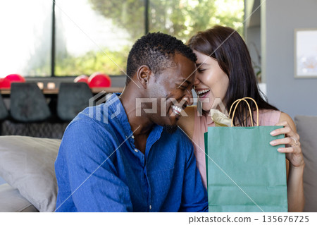 Couple laughing together while exchanging gift bag at home, enjoying special moment Couple laughing together while exchanging gift bag at home, enjoying special moment 135676725
