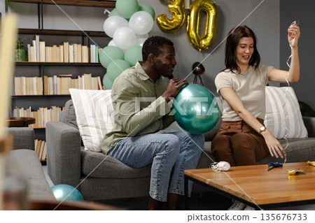 Friends preparing for birthday party, inflating balloons and decorating living room 135676733
