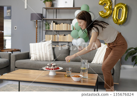 Woman arranging cupcakes on table for birthday celebration at home, smiling happily Woman arranging cupcakes on table for birthday celebration at home, smiling happily 135676824