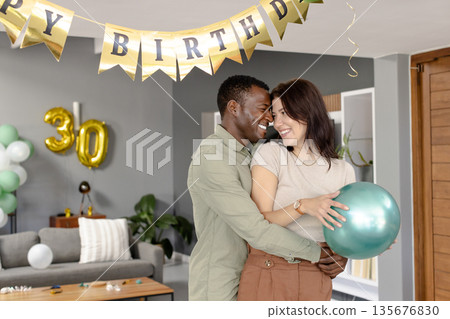 Couple celebrating birthday at home, smiling and holding balloon under decorations 135676830