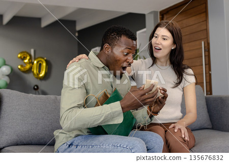 Celebrating birthday, surprised man opening gift with friend on living room sofa 135676832