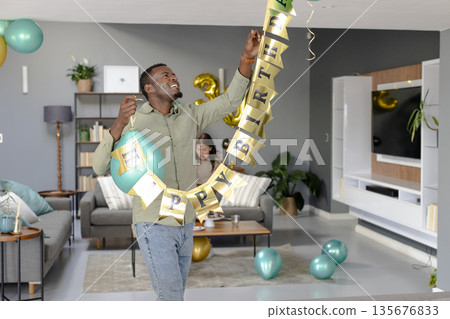 Hanging birthday banner in living room, man surrounded by balloons and smiling 135676833
