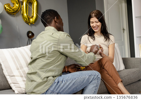 African American man proposing to smiling woman on couch during celebration African American man proposing to smiling woman on couch during celebration 135676836