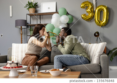Couple celebrating birthday at home, blowing party horns on couch, feeling joyful 135676848