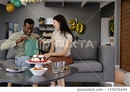 Celebrating birthday, couple exchanging gifts on sofa with cake and balloons nearby 135676849