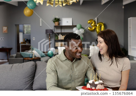 Couple celebrating birthday at home with cake and balloons, sharing joyful moment Couple celebrating birthday at home with cake and balloons, sharing joyful moment 135676864