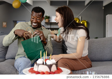 Opening gift bag, African American man and woman laughing, celebrating together on couch Opening gift bag, African American man and woman laughing, celebrating together on couch 135676866