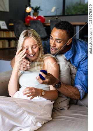 African American man proposing to surprised woman on couch at home, both smiling 135676899