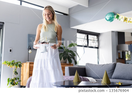 Smiling woman carrying birthday cake with candles in modern living room 135676904