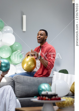 Man joyfully inflating balloons on couch during festive home celebration Man joyfully inflating balloons on couch during festive home celebration 135676910