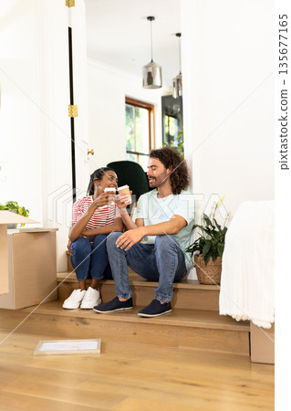 Young diverse couple sitting on stairs, enjoying coffee break while unpacking boxes at home Young diverse couple sitting on stairs, enjoying coffee break while unpacking boxes at home 135677165