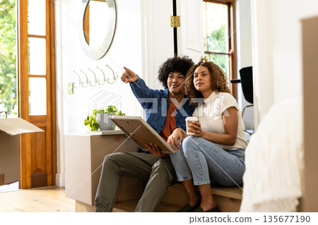 Young diverse couple planning home decor while sitting on moving boxes, feeling excited 135677190