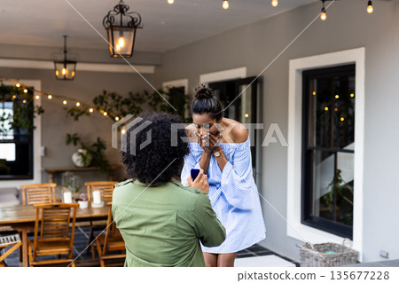 Man proposing to surprised woman on patio, romantic moment under string lights 135677228