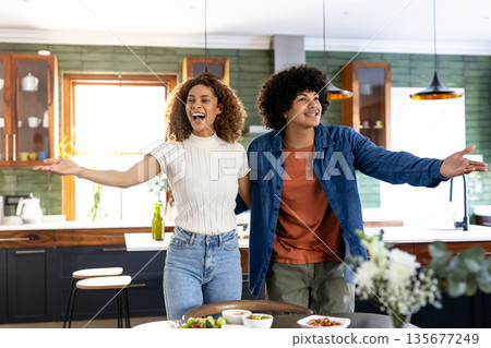 Young couple joyfully welcoming guests in modern kitchen, arms open wide 135677249