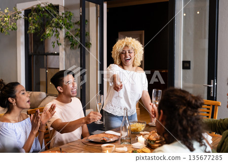 Diverse friends celebrating at dinner table, raising glasses and laughing together Diverse friends celebrating at dinner table, raising glasses and laughing together 135677285