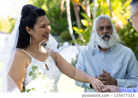 Holding hands during outdoor senior wedding, bride smiling joyfully with guests present 135677323