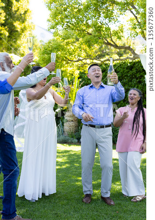 Raising champagne glasses, diverse group celebrating joyful senior wedding toast outdoors 135677360