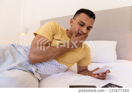 Young man lying on bed thoughtfully looking at photograph in cozy bedroom 135677369