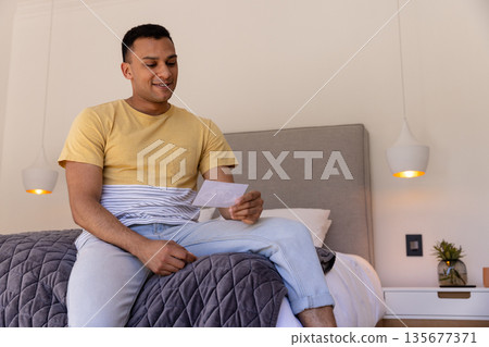 Young man sitting on bed holding photo, smiling and reminiscing at home 135677371