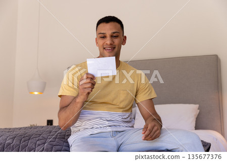 Young man sitting on bed holding photograph, smiling with joy and nostalgia Young man sitting on bed holding photograph, smiling with joy and nostalgia 135677376