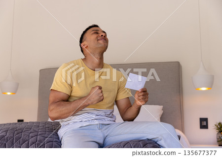 Young man sitting on bed celebrating while holding photograph, feeling joyful Young man sitting on bed celebrating while holding photograph, feeling joyful 135677377