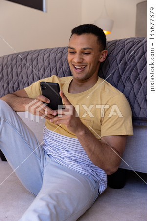 Smiling young man using smartphone while relaxing on floor at home Smiling young man using smartphone while relaxing on floor at home 135677379