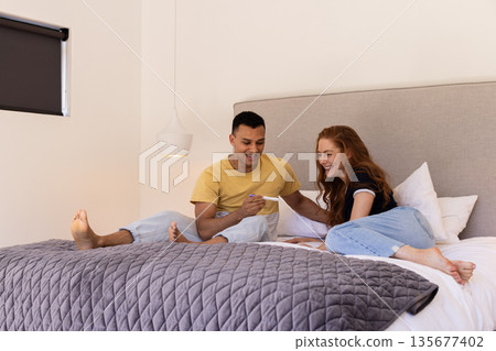 Young couple sitting on bed, joyfully looking at pregnancy test together 135677402